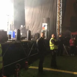 Ochrona imprezy masowej: ochroniarze w kamizelkach odblaskowych stoją przed sceną podczas koncertu plenerowego w nocy.