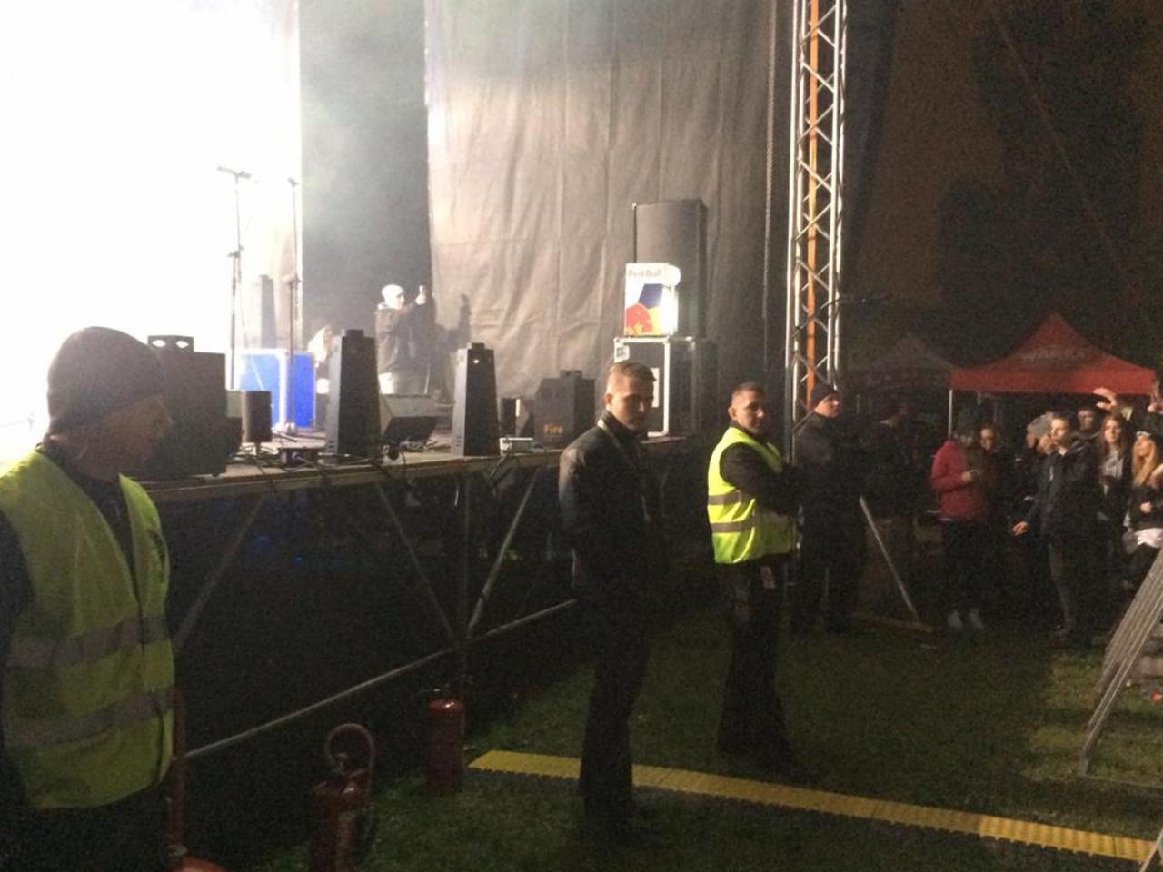 Ochrona imprezy masowej: ochroniarze w kamizelkach odblaskowych stoją przed sceną podczas koncertu plenerowego w nocy.