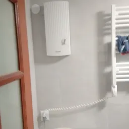 Biały, elektryczny podgrzewacz wody marki Stiebel Eltron zamontowany na szarej, kafelkowanej ścianie w łazience, obok grzejnika drabinkowego z powieszonym ręcznikiem, widoczny fragment umywalki...