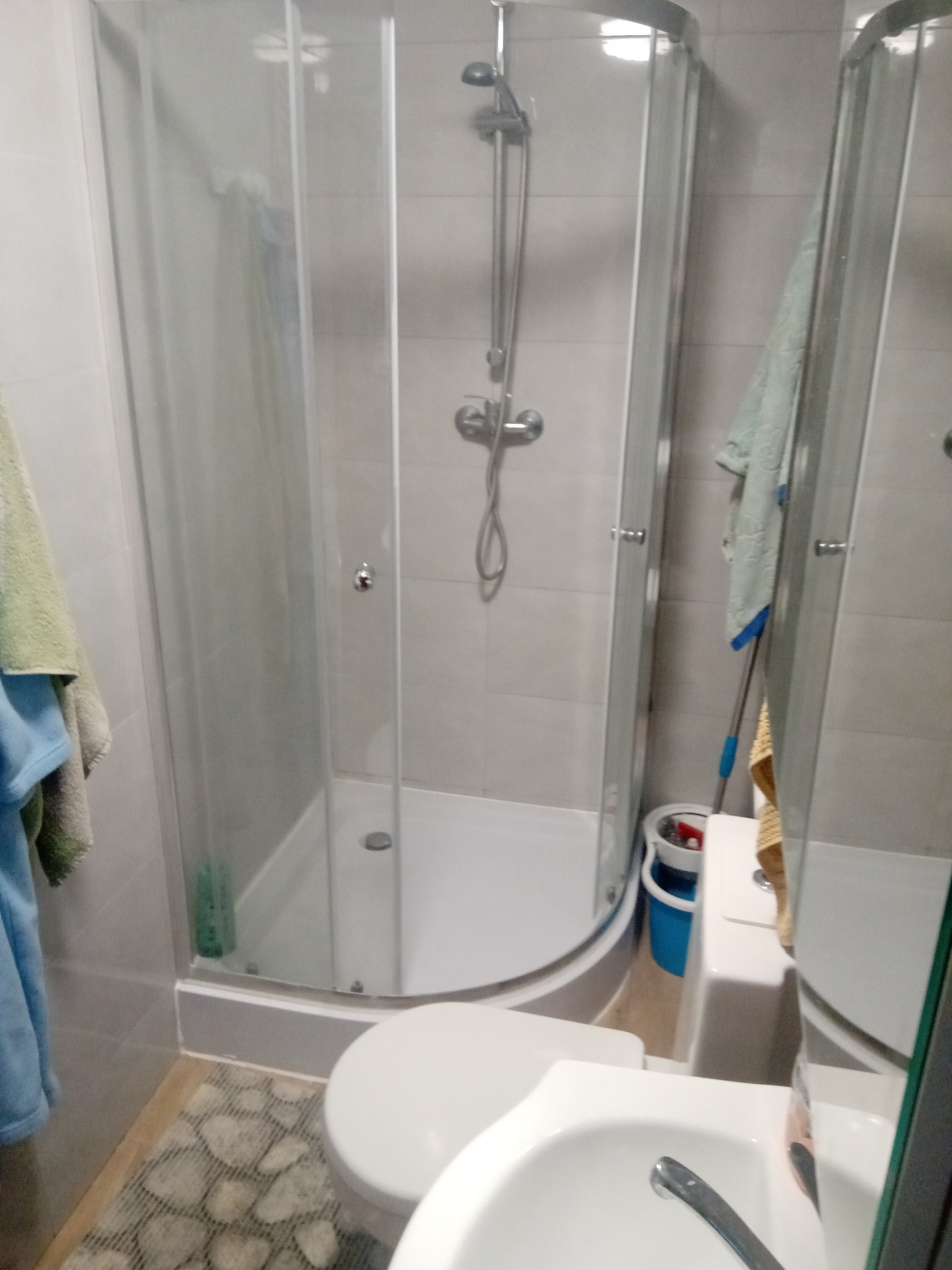 Wnętrze łazienki z kabiną prysznicową z zaokrąglonymi szybami, widok na białą umywalkę, toaletę oraz szary dywanik imitujący kamienie.