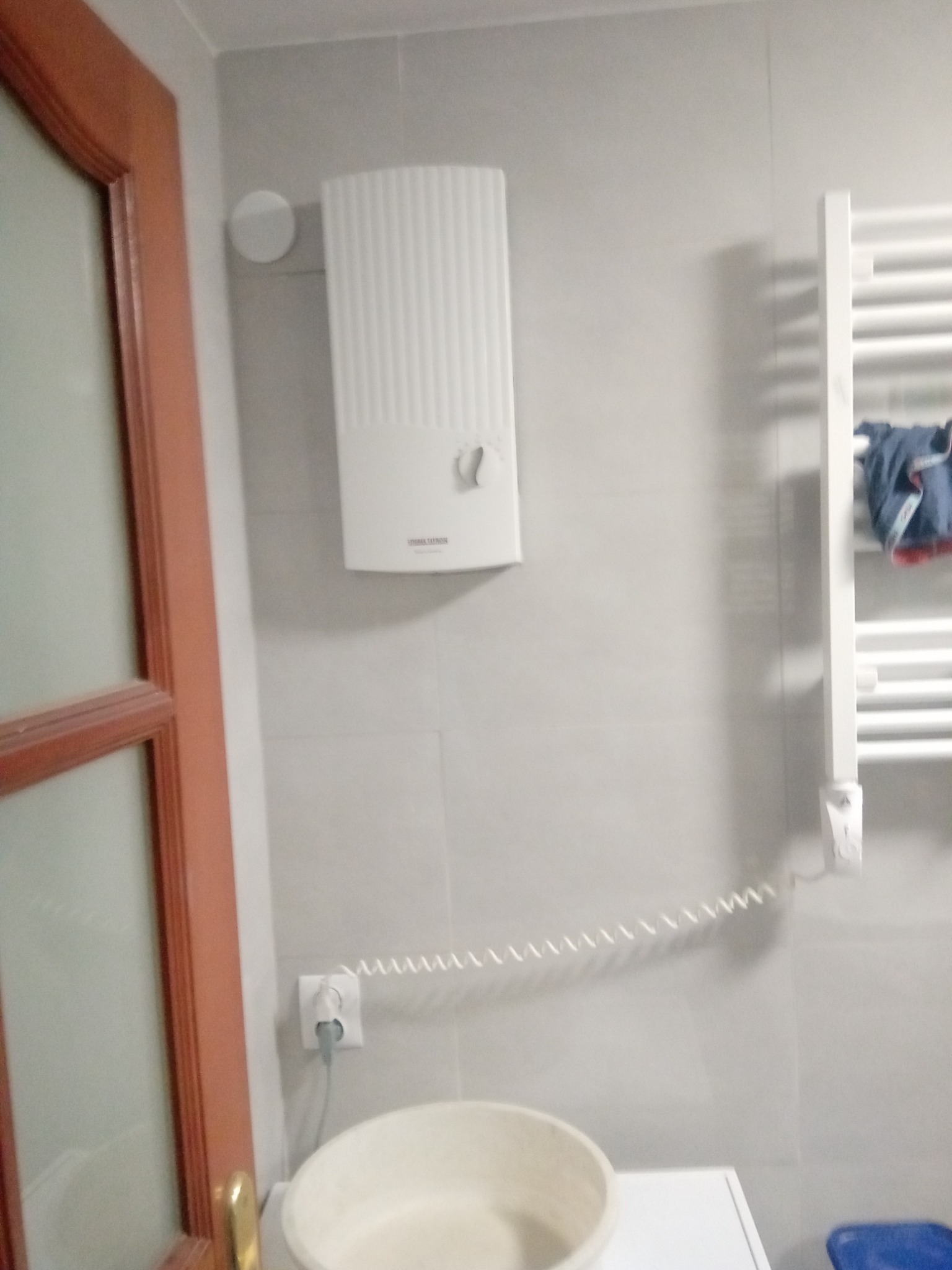 Biały, elektryczny podgrzewacz wody marki Stiebel Eltron zamontowany na szarej, kafelkowanej ścianie w łazience, obok grzejnika drabinkowego z powieszonym ręcznikiem, widoczny fragment umywalki...