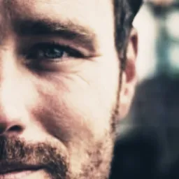 Portret mężczyzny z brodą, widoczna prawa strona twarzy, błękitne oko, ciemne tło.