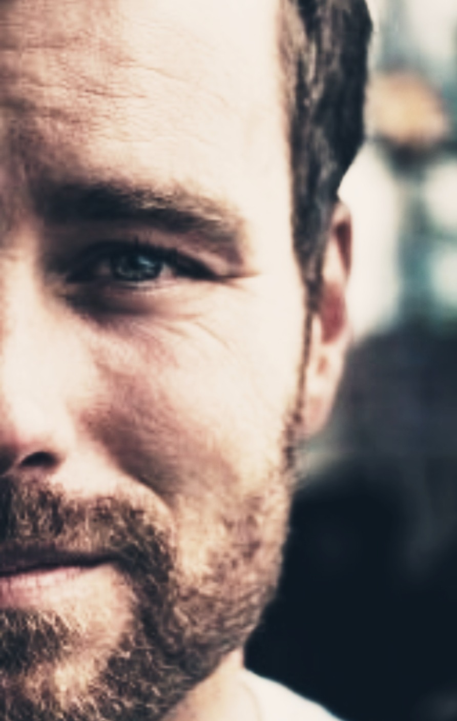 Portret mężczyzny z brodą, widoczna prawa strona twarzy, błękitne oko, ciemne tło.