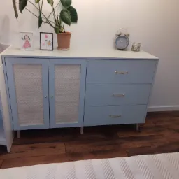 Niebieska komoda z trzema szufladami i dwojgiem drzwi z rattanowym wypełnieniem, ustawiona na drewnianej podłodze, z dekoracjami na blacie, w pokoju dziecięcym.