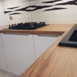 Fragment blatu kuchennego z drewna o jasnym odcieniu, z zamontowaną czarną płytą grzewczą i zlewozmywakiem, na tle białych szafek i płytek z wzorem zebry.