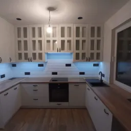 Jasna kuchnia z drewnianymi blatami i białymi szafkami z przeszklonymi frontami, czarna armatura i płytki imitujące cegłę z ciemnymi fugami, oświetlenie podszafkowe.