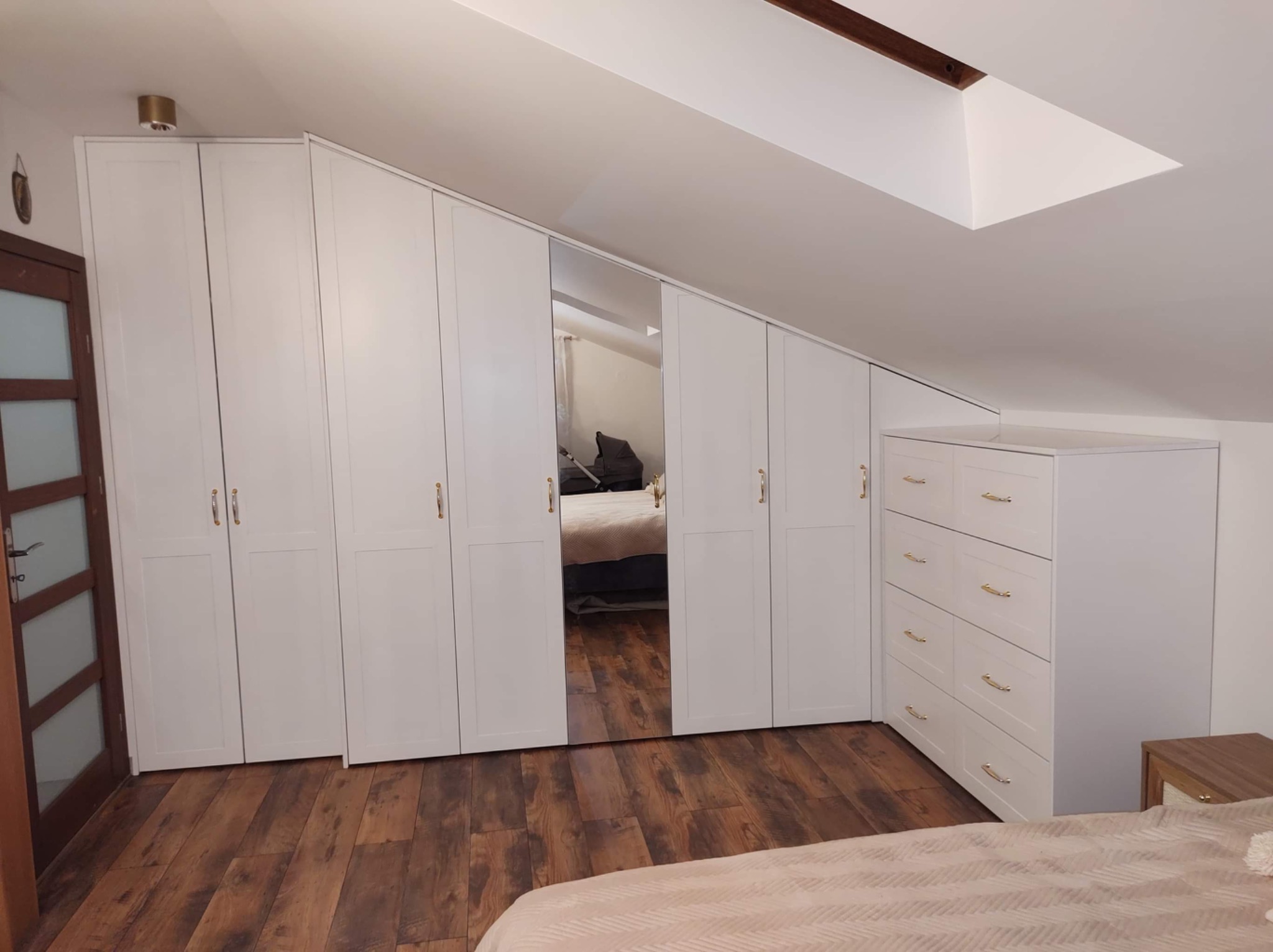 Biała szafa w zabudowie na poddaszu ze skośnym sufitem, z lustrzanymi drzwiami i komodą z szufladami, drewniana podłoga, widok na łóżko w odbiciu lustra.