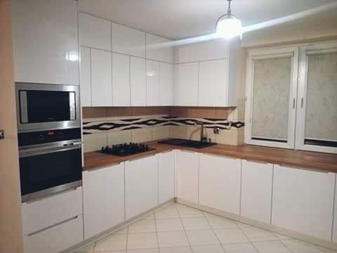 Nowoczesna kuchnia z białymi, lakierowanymi frontami, drewnianym blatem i płytkami z geometrycznym wzorem na ścianie, widoczna zabudowa piekarnika i płyta gazowa.