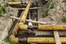 Rury w żółtej izolacji termicznej ułożone w wykopie ziemnym, zabezpieczone deskami.