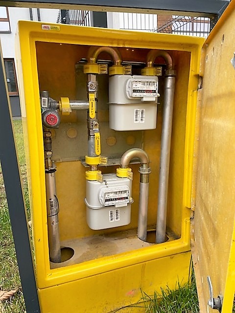 Dwa gazomierze w żółtej skrzynce przyłączeniowej, widoczne rury i zawory, trawa u podstawy skrzynki.