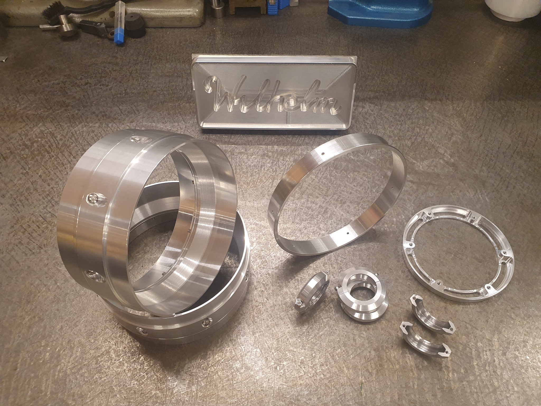 Precyzyjnie obrobione, błyszczące, metalowe pierścienie i elementy z otworami na szorstkiej powierzchni warsztatowej, obok wyfrezowanego bloku z napisem 'Wilhelm', narzędzia w tle.