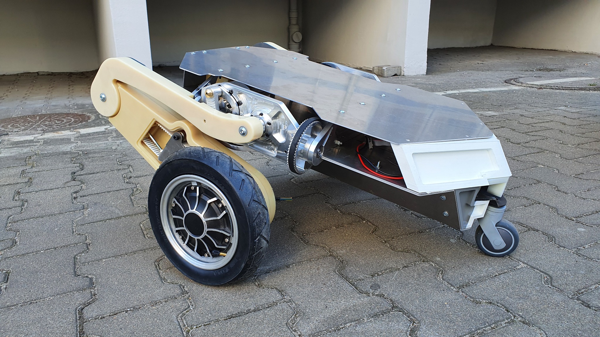 Prototyp pojazdu elektrycznego z widocznymi elementami konstrukcyjnymi, w tym kołem z silnikiem, przekładnią pasową i elementami z tworzywa sztucznego, na tle brukowanej nawierzchni.