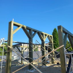 Firma Remontowo-Budowlana Pilch Robert - Konstrukcja drewniana w trakcie budowy, zielone belki połączone w szkielet, widoczne rusztowania i narzędzia, tło: błękitne niebo i budynek w budowie.