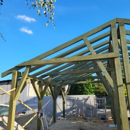 Firma Remontowo-Budowlana Pilch Robert - Konstrukcja drewniana z zielonych belek na żwirowym podłożu, w tle ściana i rusztowania pod błękitnym niebem z liśćmi. Etap budowy altany lub wiaty.