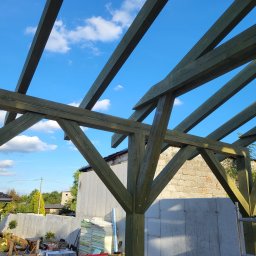 Firma Remontowo-Budowlana Pilch Robert - Konstrukcja drewniana z belek w kolorze zielonym, widok z dołu na błękitne niebo z chmurami. W tle budynek z betonu i cegły, materiały budowlane i narzędzia.