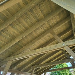 Firma Remontowo-Budowlana Pilch Robert - Drewniana konstrukcja dachu altany w trakcie budowy, widok z dołu, ukazujący belkowanie i deskowanie pomalowane na zielono-brązowy kolor, z elementami krajobrazu w tle.