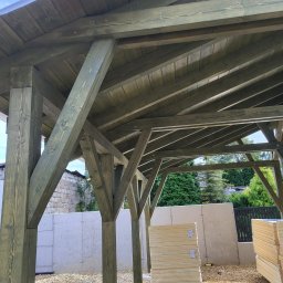 Firma Remontowo-Budowlana Pilch Robert - Drewniana konstrukcja z widocznym dachem i belkami, w tle zaplecze budowy z izolacją i taczką, ujęcie od dołu, ukazujące detale konstrukcyjne.