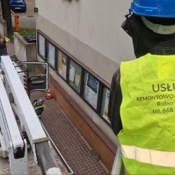 Firma Remontowo-Budowlana Pilch Robert - Pracownik w żółtej kamizelce z napisem 'Usługi Remontowo-Budowlane' na podnośniku koszowym podczas prac przy elewacji budynku z plakatami.
