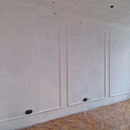 Firma Remontowo-Budowlana Pilch Robert - Białe ściany z dekoracyjnymi listwami i gniazdkami, podłoga z parkietu w jodełkę. Widoczne detale wykończenia wnętrza po malowaniu. Oświetlenie sufitowe punktowe.