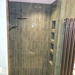 Firma Remontowo-Budowlana Pilch Robert - Wykończona kabina prysznicowa z zielonymi, wąskimi płytkami ułożonymi pionowo, wbudowanymi półkami i czarną armaturą. Widoczny fragment grzejnika.