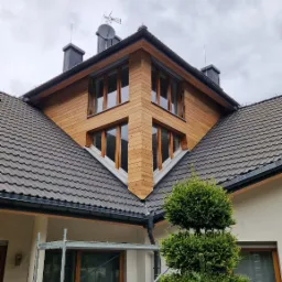 Elewacja domu z drewnianą okładziną, widok z dołu na narożnik budynku. Część elewacji zakryta rusztowaniem i krzewem.