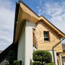 Nowoczesny dom z drewnianą elewacją i białym elementem architektonicznym, widok z dołu na tle błękitnego nieba.