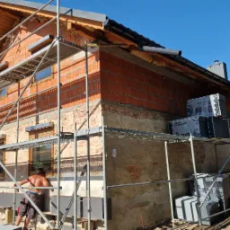 Budowa domu z cegły: murarz przy rusztowaniu ociepla ścianę szarą płytą izolacyjną. Częściowo otynkowana elewacja, widoczne pakiety izolacyjne i błękitne niebo.