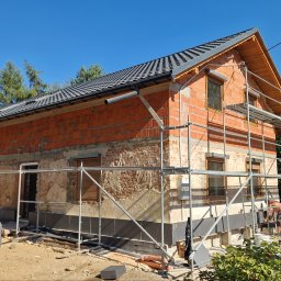 Firma Remontowo-Budowlana Pilch Robert - Dom w trakcie prac elewacyjnych: widoczne rusztowania, fragmenty ocieplenia, ceglany mur i nowa dachówka na tle błękitnego nieba.