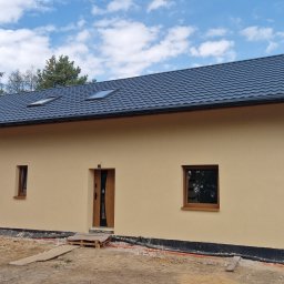 Firma Remontowo-Budowlana Pilch Robert - Nowoczesny dom jednorodzinny z beżową elewacją i ciemnym, metalowym dachem. Widoczne okna dachowe i elementy wykończeniowe. Wokół domu teren w trakcie prac ziemnych.