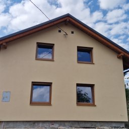 Firma Remontowo-Budowlana Pilch Robert - Dwukondygnacyjny dom z beżową elewacją i drewnianymi ramami okiennymi w słoneczny dzień z widocznymi elementami wykończeniowymi dachu i instalacją elektryczną.