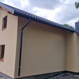 Dwukondygnacyjny dom z beżową elewacją i ciemnymi rynnami, widoczny z boku. Dach pokryty czarną dachówką. Okna z brązowymi ramami. Częściowo widoczne drzewa w tle.