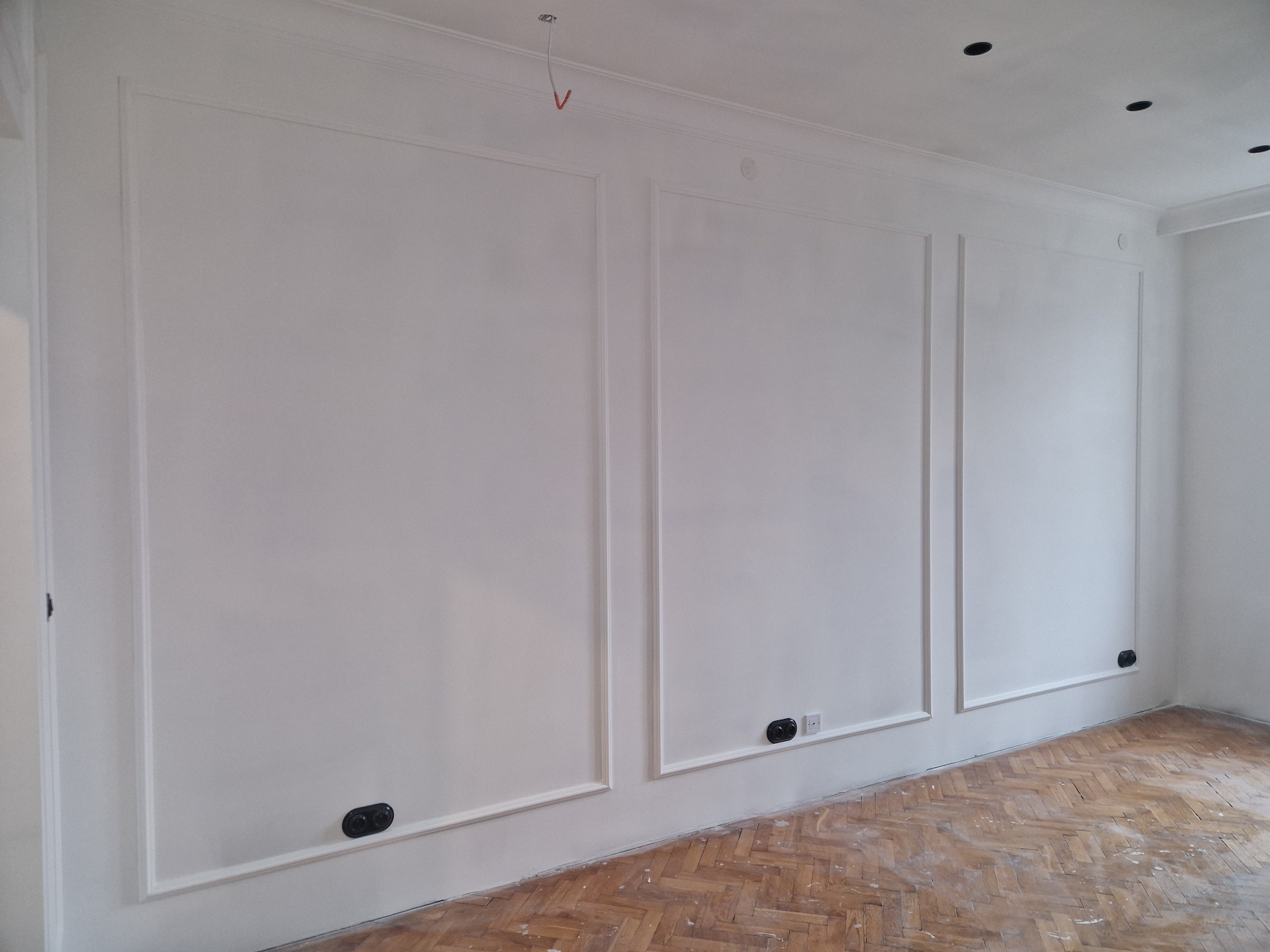 Białe ściany z dekoracyjnymi listwami i gniazdkami, podłoga z parkietu w jodełkę. Widoczne detale wykończenia wnętrza po malowaniu. Oświetlenie sufitowe punktowe.