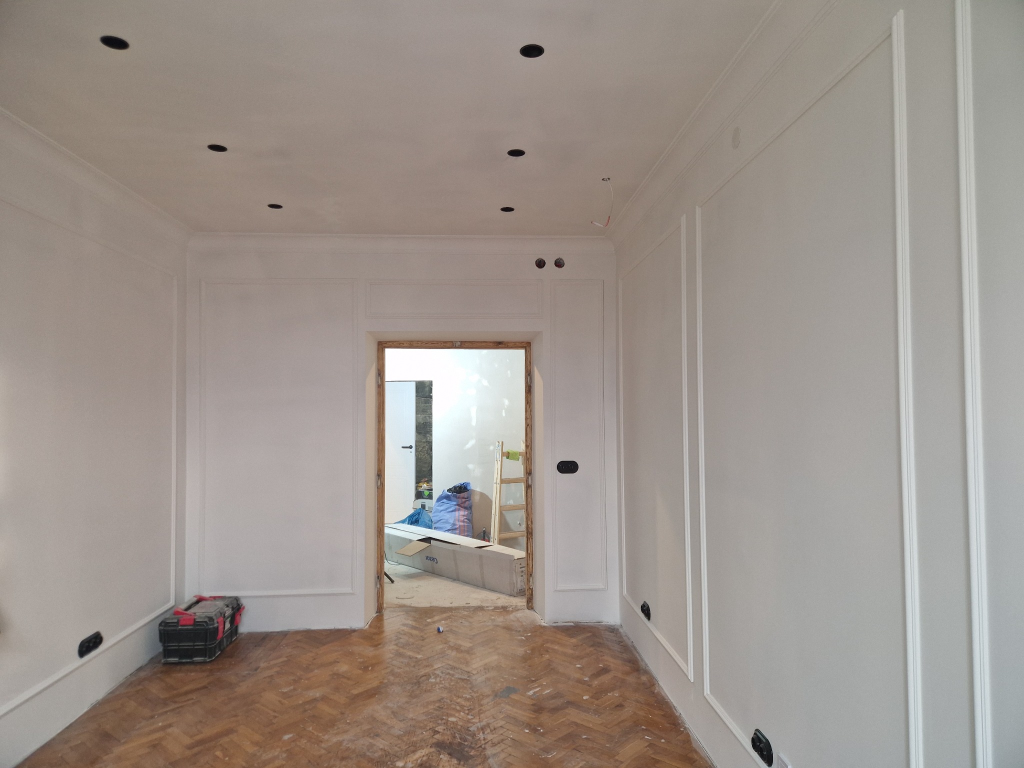Białe ściany z dekoracyjnymi listwami i parkietem w jodełkę. Widoczne otwory na oświetlenie w suficie i skrzynka narzędziowa w rogu. W tle widok na kolejne pomieszczenie.