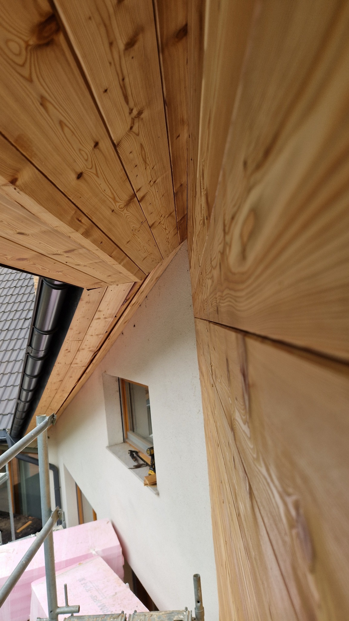 Montaż drewnianej elewacji na szczycie domu, widoczne rusztowanie, okno i narzędzia. Ujęcie z dołu, ukazujące detale drewnianej konstrukcji i ocieplenie.