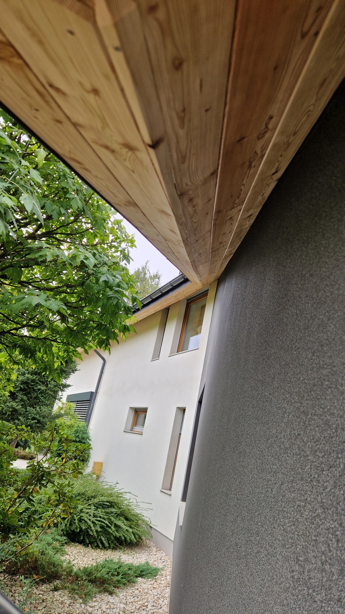 Nowoczesny dom z drewnianą podbitką dachową i elewacją w dwóch odcieniach szarości, otoczony zielenią. Ujęcie z dołu podkreśla geometrię budynku.