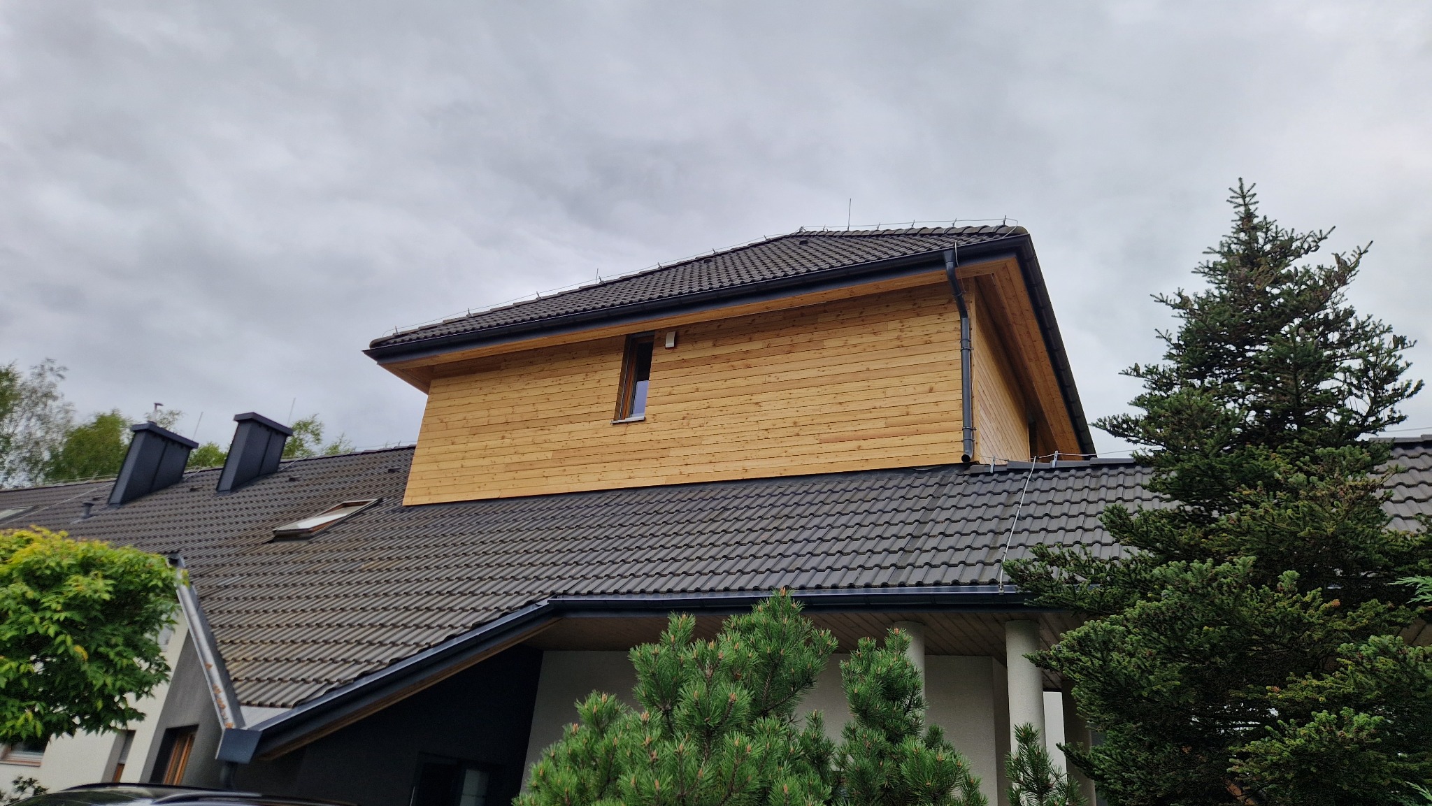Dwukondygnacyjny dom z drewnianą elewacją i ciemnym dachem z dachówki, widziany z dołu, z zielenią w tle i pochmurnym niebem.