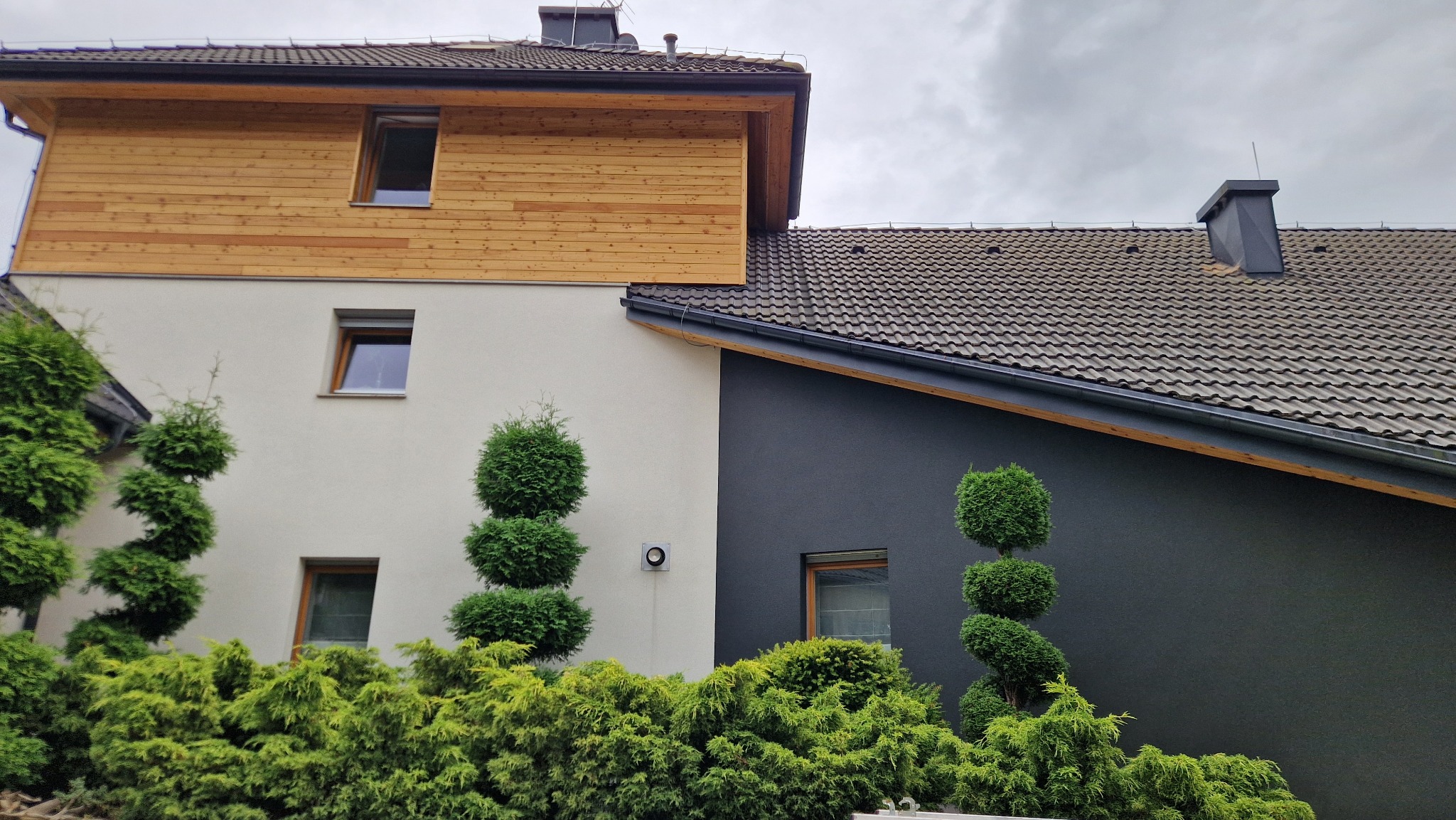 Dwukondygnacyjny dom z elewacją w kolorach: biały, grafitowy i drewno. Z przodu ozdobne krzewy. Dachówka ceramiczna. Komin z blachy w kolorze grafitu.