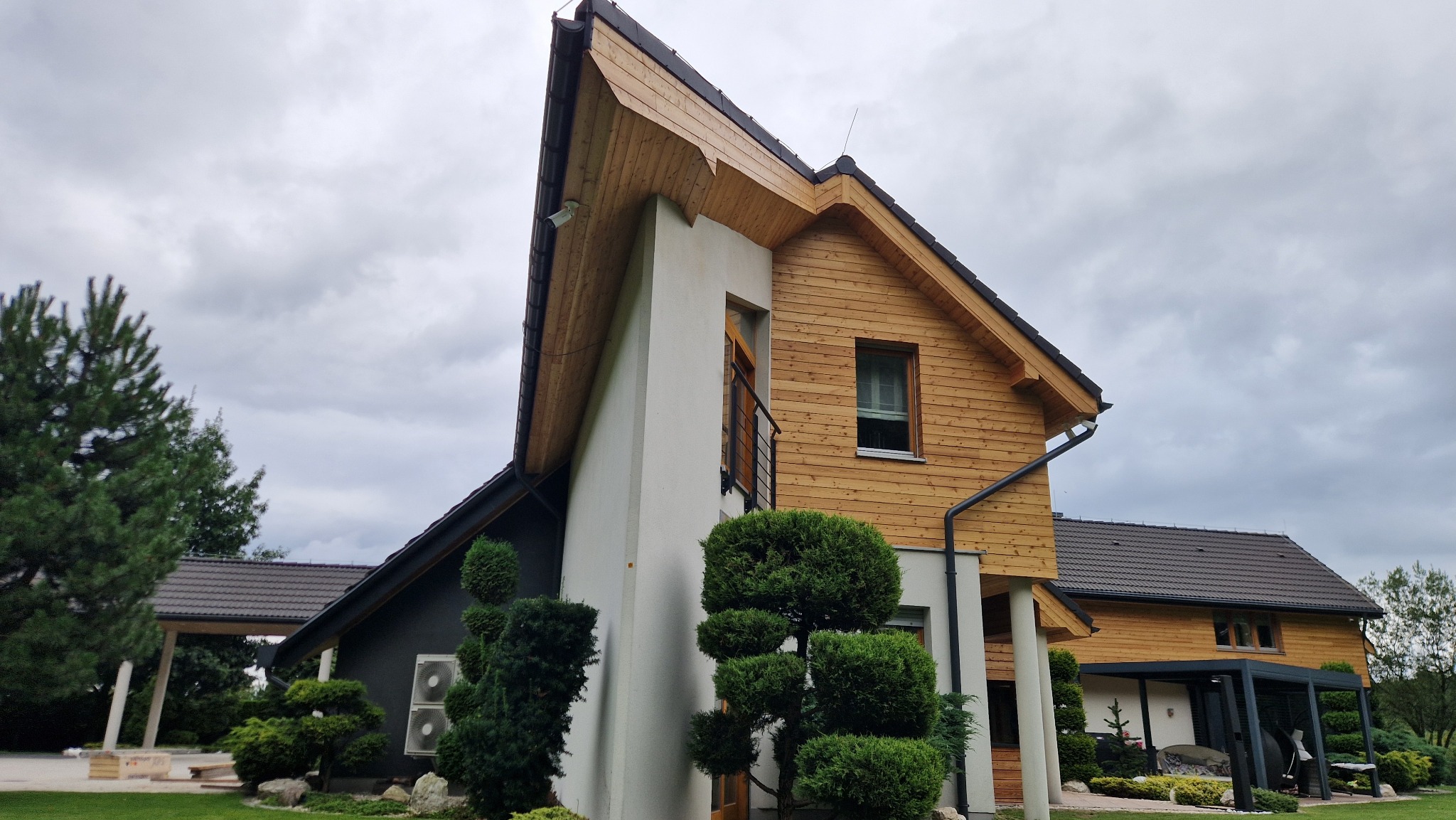 Nowoczesny dom z drewnianą elewacją i białymi elementami, otoczony zadbanym ogrodem, na tle pochmurnego nieba. Widoczny fragment dachu i elementy wykończeniowe.