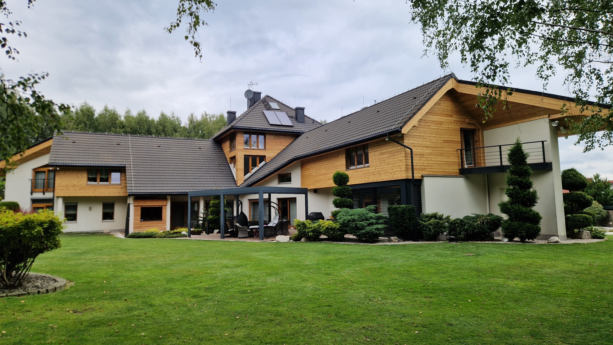 Nowoczesny dom z drewnianą elewacją i szarą dachówką, otoczony zielonym trawnikiem i ozdobnymi krzewami. Widoczne panele solarne na dachu.