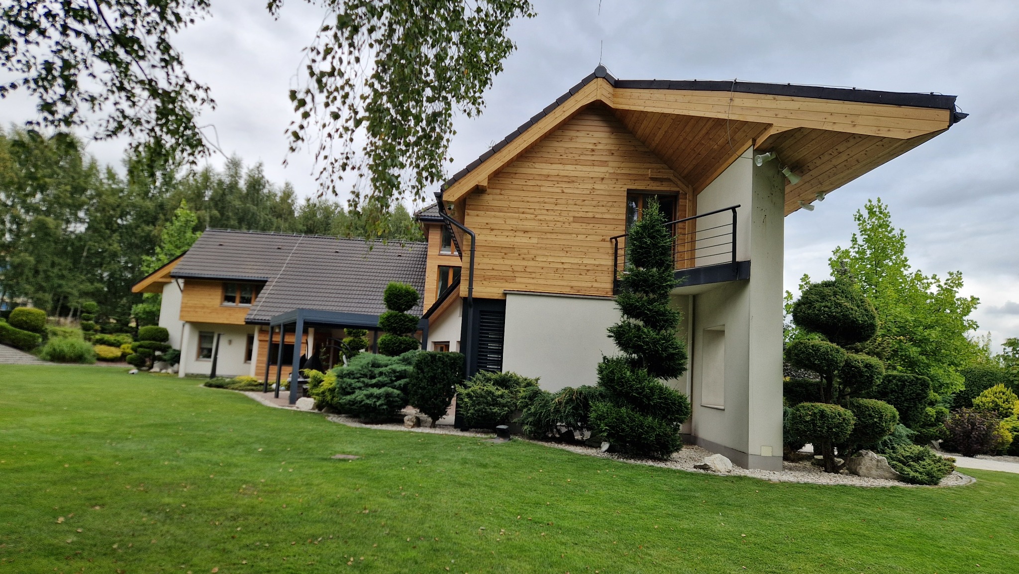 Nowoczesny dom z drewnianą elewacją i balkonem, otoczony zielenią. Widoczne starannie przystrzyżone krzewy i trawnik, tworzące harmonijną całość.