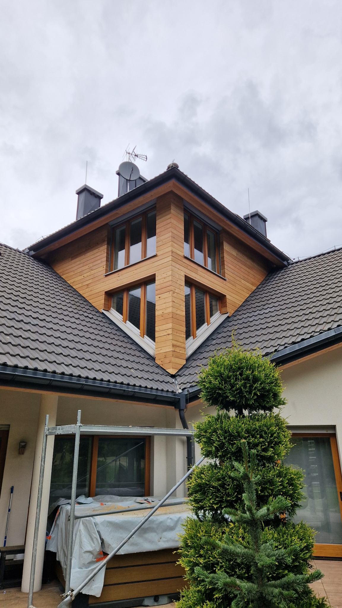 Elewacja domu z drewnianą okładziną, widok z dołu na narożnik budynku. Część elewacji zakryta rusztowaniem i krzewem.