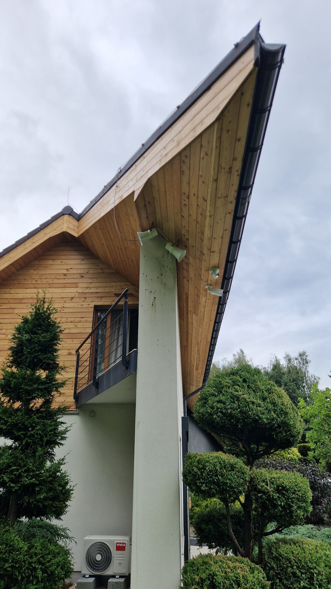 Nowoczesny dom z drewnianą elewacją i nietypowym dachem. Widoczne oświetlenie zewnętrzne, balkon z czarną balustradą i klimatyzator. Zieleń w tle.
