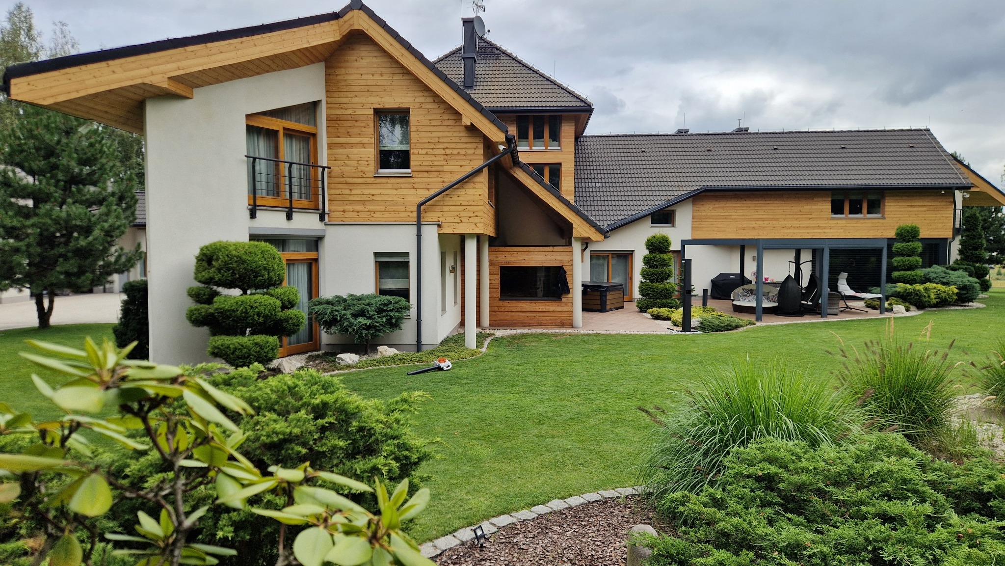 Nowoczesny dom z elementami drewnianymi na elewacji, zadbany trawnik i dekoracyjne krzewy w ogrodzie. Widoczny fragment tarasu z meblami ogrodowymi.