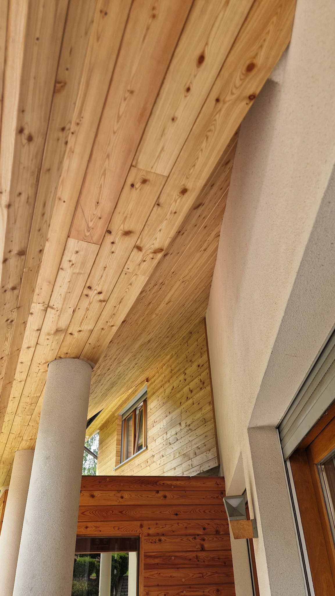 Drewniany sufit z widoczną fakturą słojów, wsparty na kolumnach, z fragmentem elewacji budynku w tle, widziany z dołu pod kątem. Naturalne kolory drewna i jasny odcień elewacji.