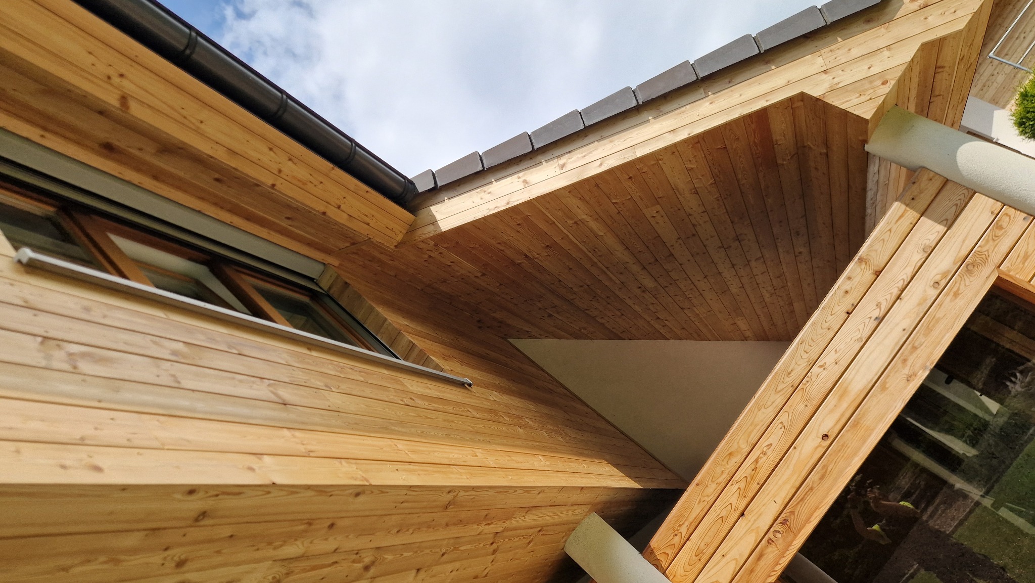 Nowoczesny dom z elewacją z jasnego drewna, widok z dołu ukazujący okno, rynnę i fragment dachu na tle błękitnego nieba. Drewniana boazeria na zewnątrz budynku.