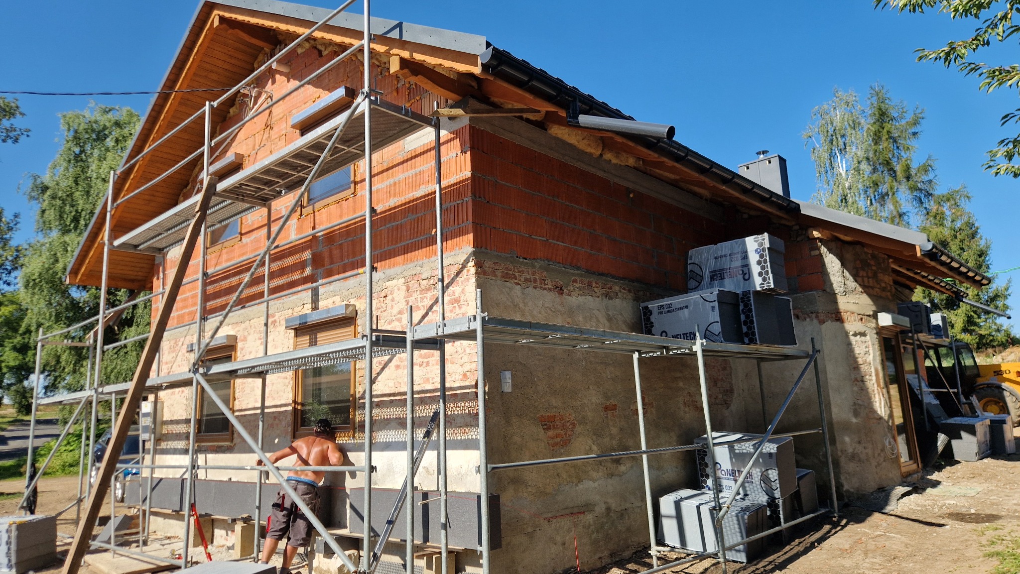 Budowa domu z cegły: murarz przy rusztowaniu ociepla ścianę szarą płytą izolacyjną. Częściowo otynkowana elewacja, widoczne pakiety izolacyjne i błękitne niebo.