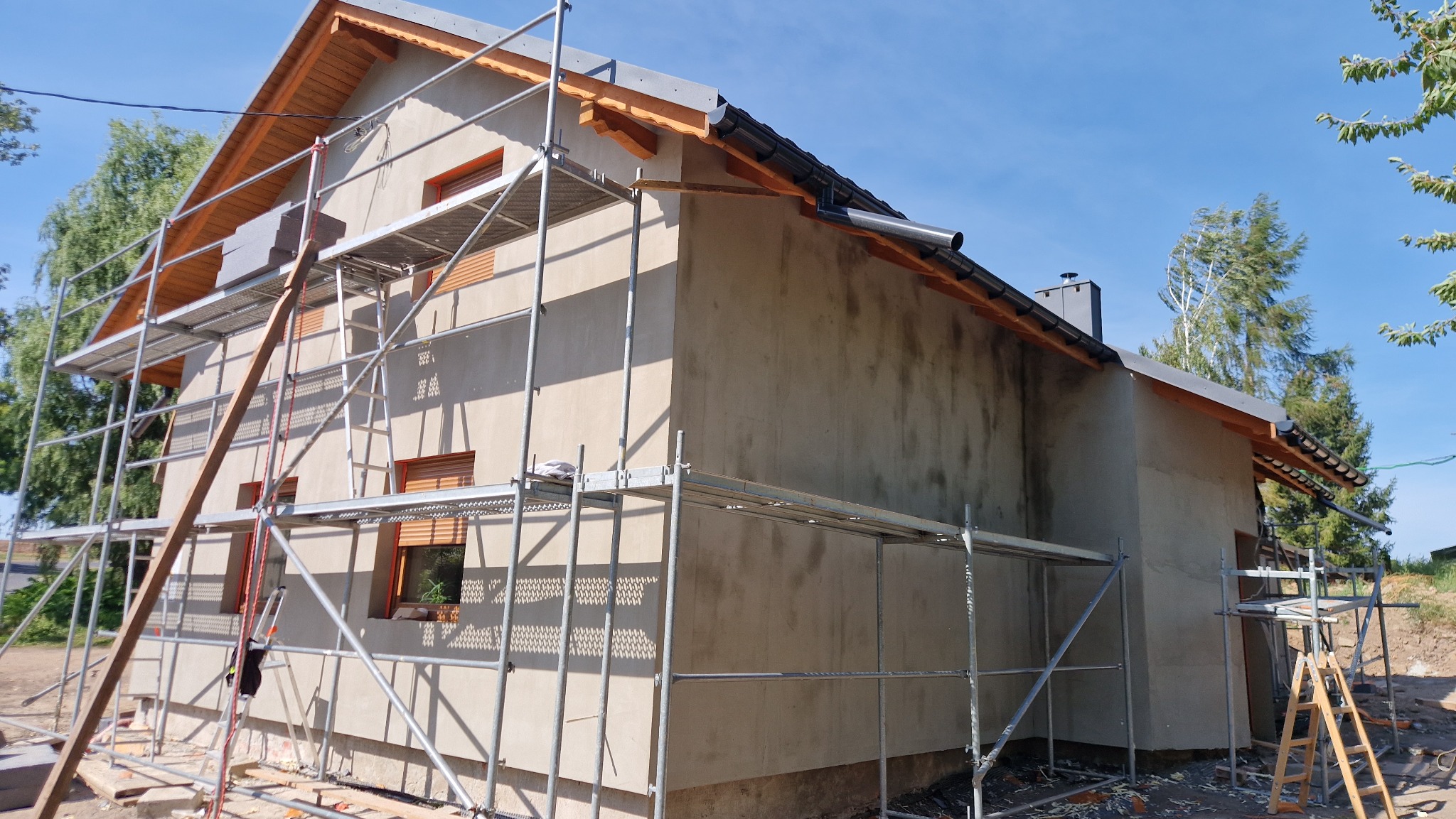 Dom w trakcie ocieplania, widoczne rusztowania i surowa elewacja. Dach pokryty dachówką, okna z brązowymi ramami. Jasne niebo w tle.