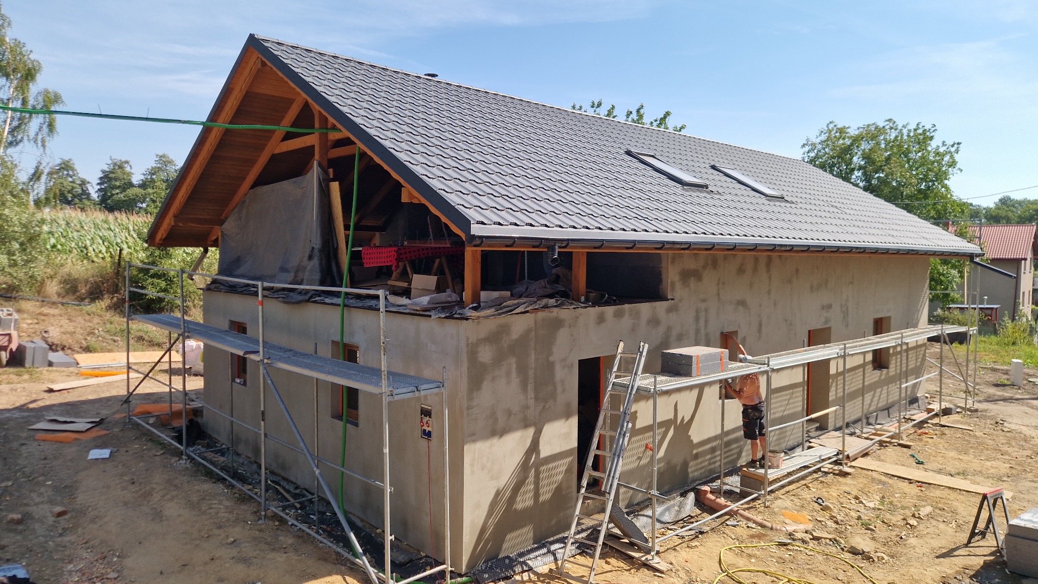 Budowa domu w trakcie: surowa elewacja, rusztowania, częściowo ukończony dach z oknami. Widoczny pracownik na rusztowaniu i otoczenie z zielenią.