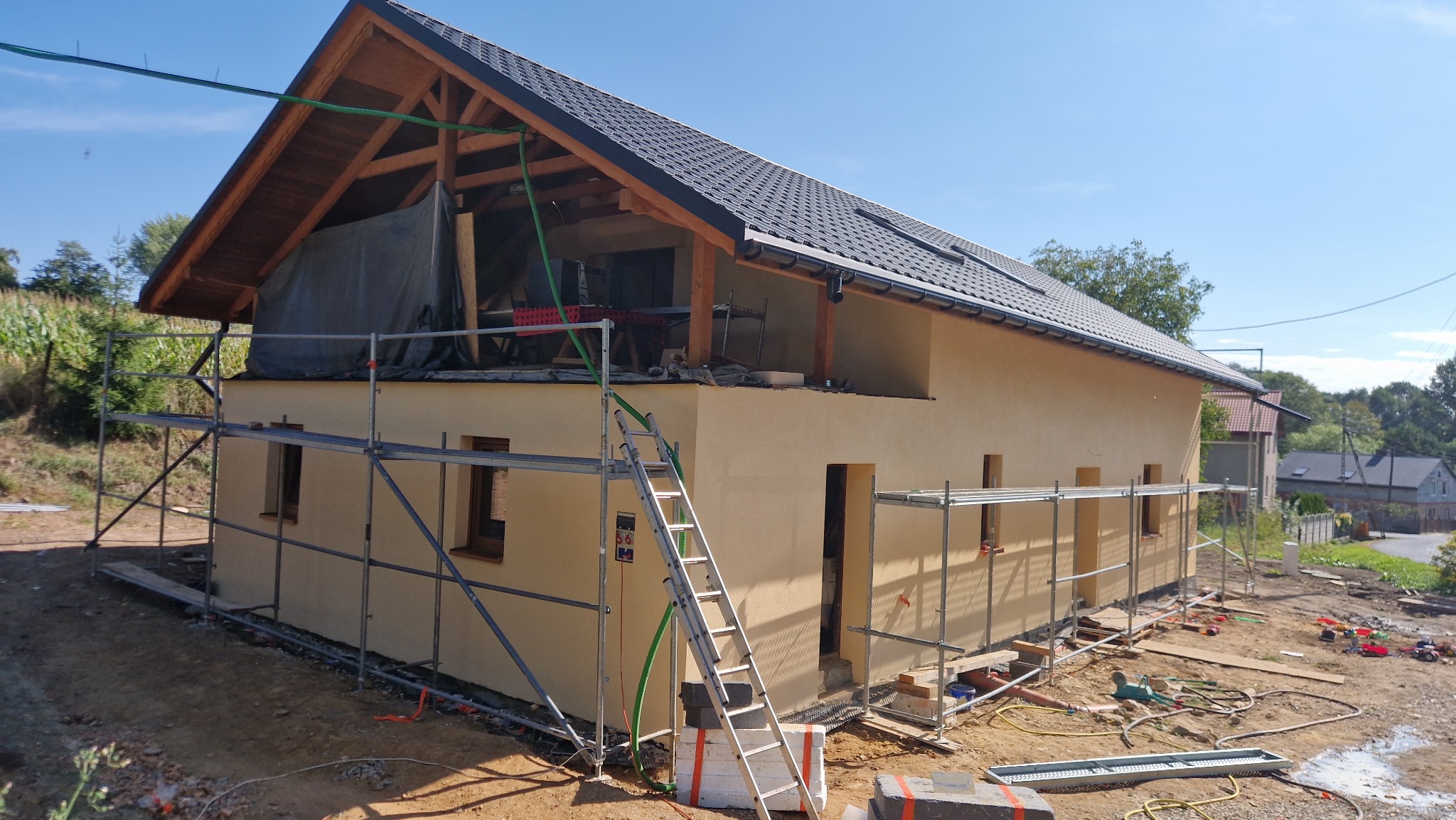 Dom w trakcie budowy z widocznym rusztowaniem, elewacją w kolorze beżowym i dachem z czarnej dachówki. Widoczne elementy drewniane konstrukcji dachu.