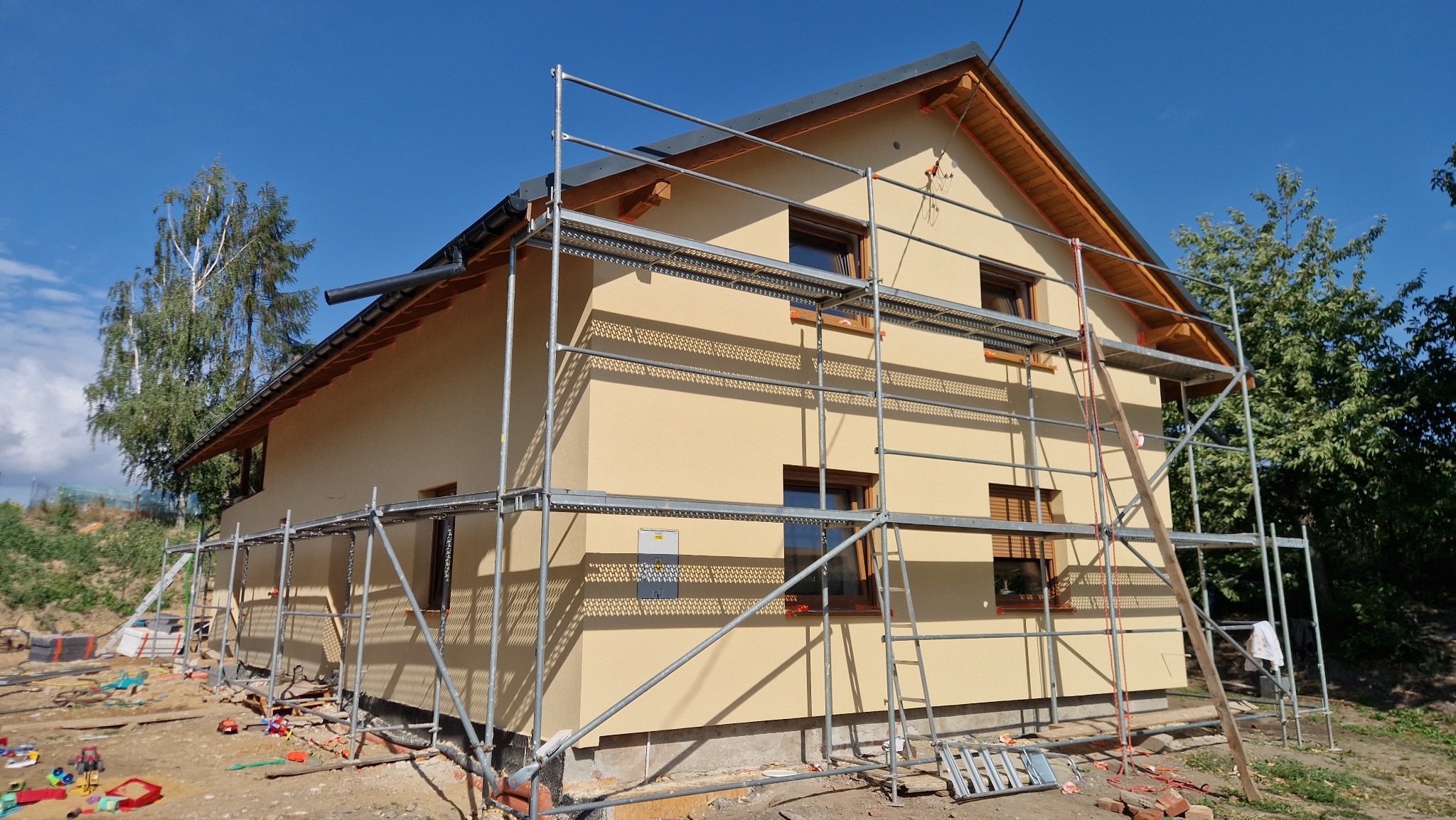 Nowo otynkowany dom jednorodzinny w kolorze beżowym, otoczony rusztowaniem i częściowo zagospodarowanym terenem. Widoczna drabina oparta o ścianę.